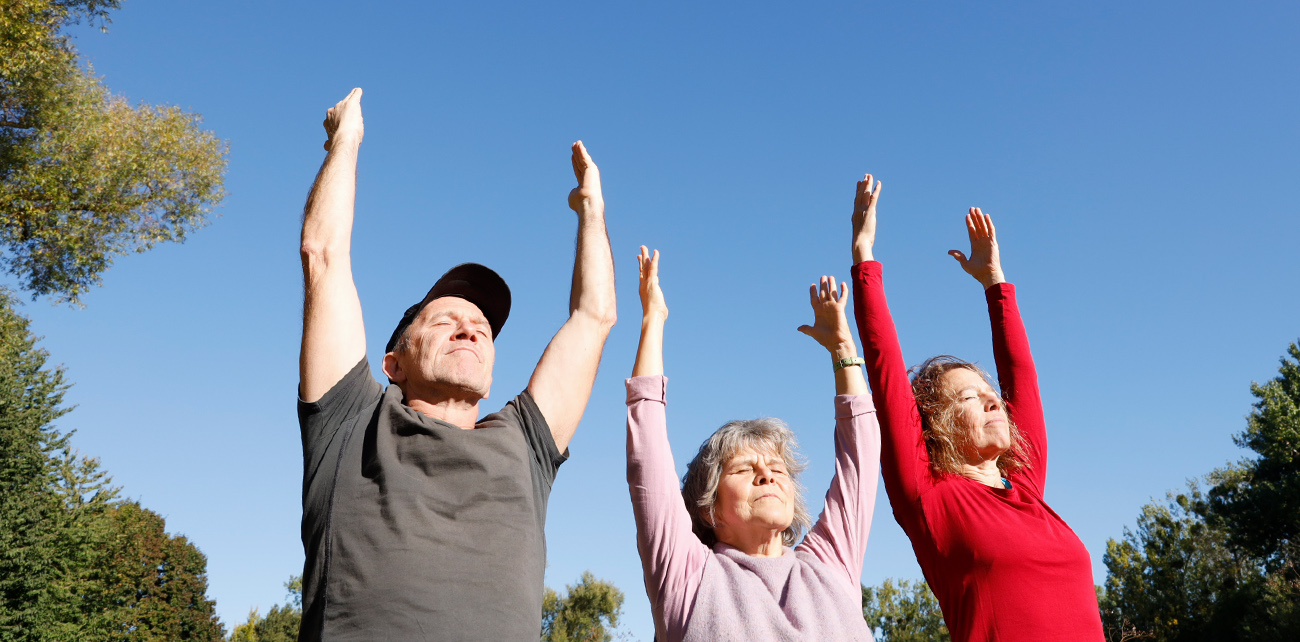 Ein Mann und zwei Frauen mittleren Alters, gestreckte Arme nach oben vor blauem Himmel, nur Oberkörper