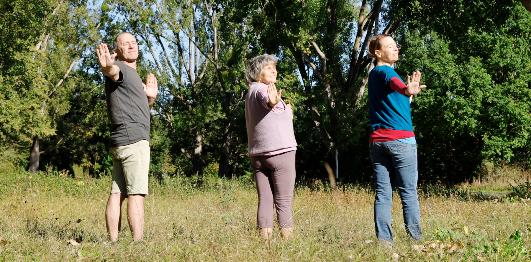 Ein Mann und zwei Frauen stehen auf einer Wiese, machen Entspannungsübungen